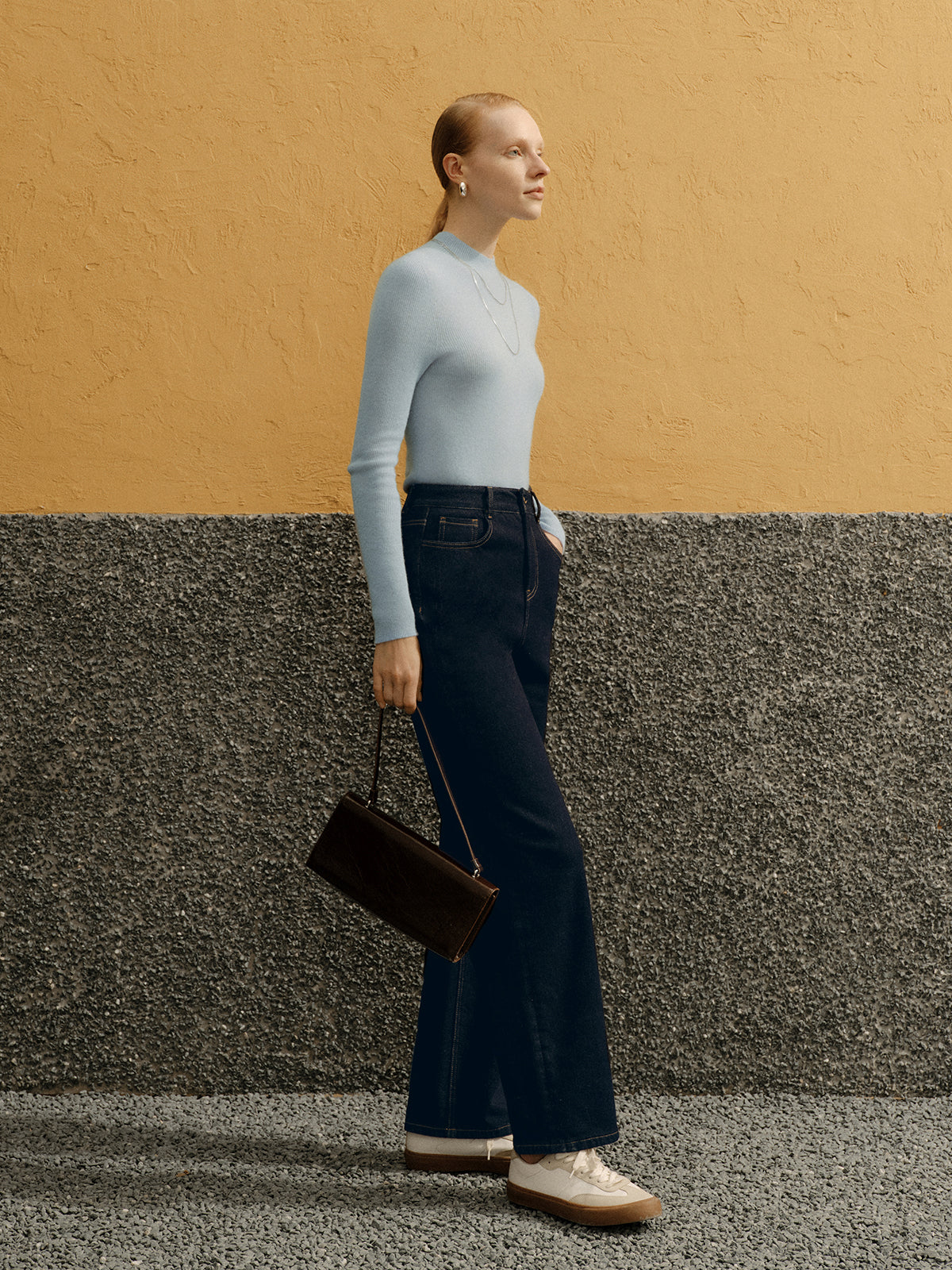Woman in a light gray turtleneck and dark blue jeans standing against a yellow and gray wall.