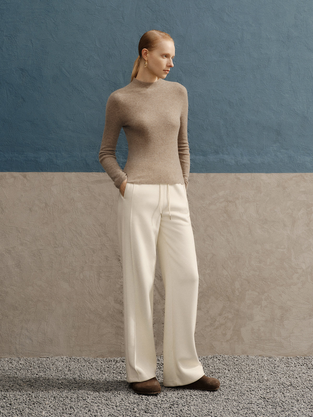 Woman wearing a beige sweater and white pants against a blue wall.
