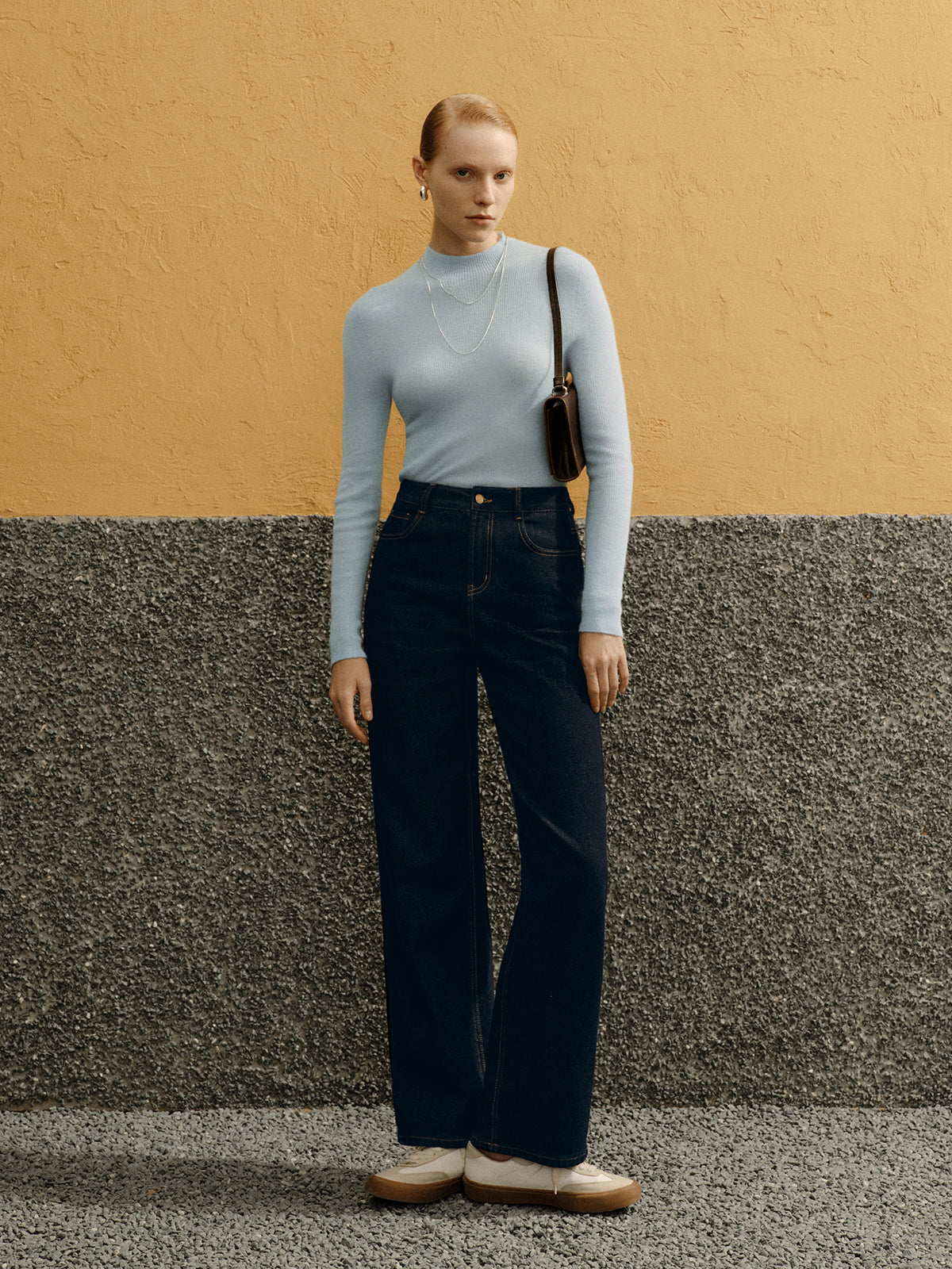 Woman in light blue top and dark pants standing against a textured wall.