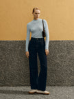 Woman in light blue top and dark pants standing against a textured wall.