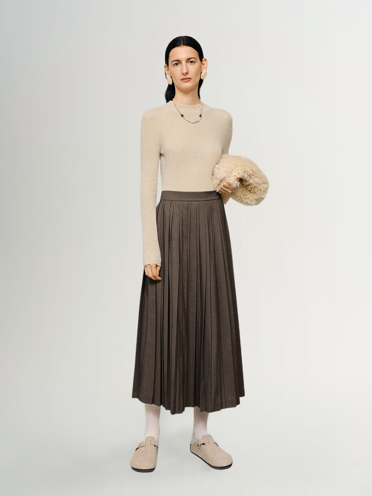 Woman wearing a beige sweater and brown pleated skirt holding a beige bag on a white background