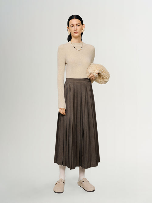 Load image 1 in gallery view. Woman wearing a beige sweater and brown pleated skirt holding a beige bag on a white background