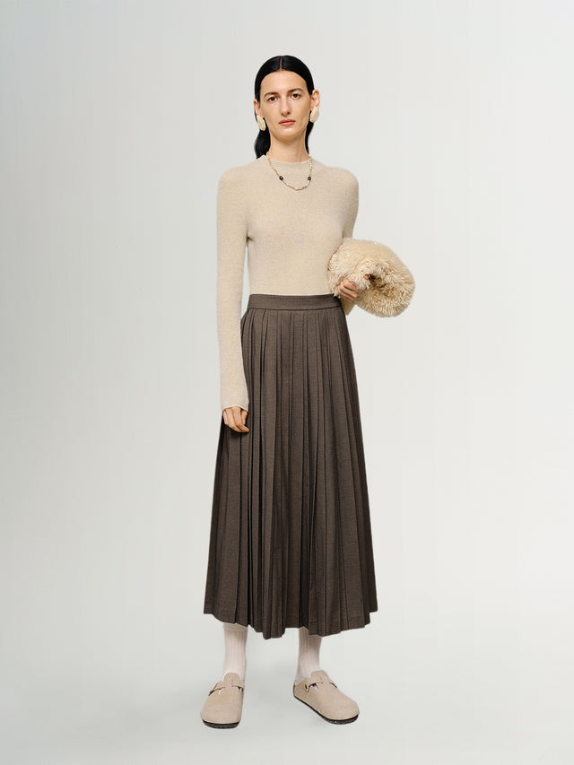 Woman wearing a beige sweater and brown pleated skirt holding a beige bag on a white background