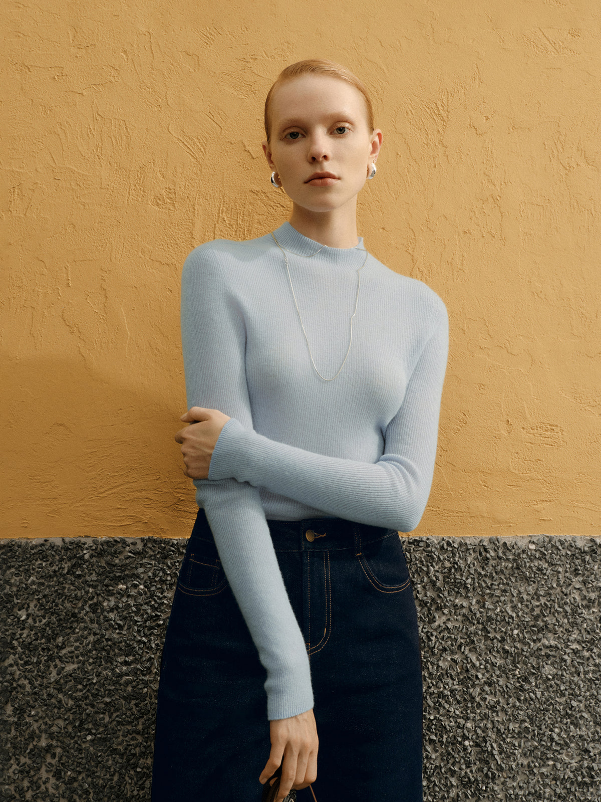 Woman wearing a light blue sweater and dark jeans against a textured beige wall.